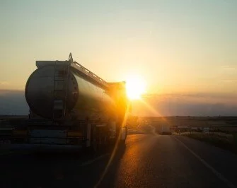 Tanker truck driving along a highway at sunset, with warm sunlight reflecting off the trailer and the road ahead.